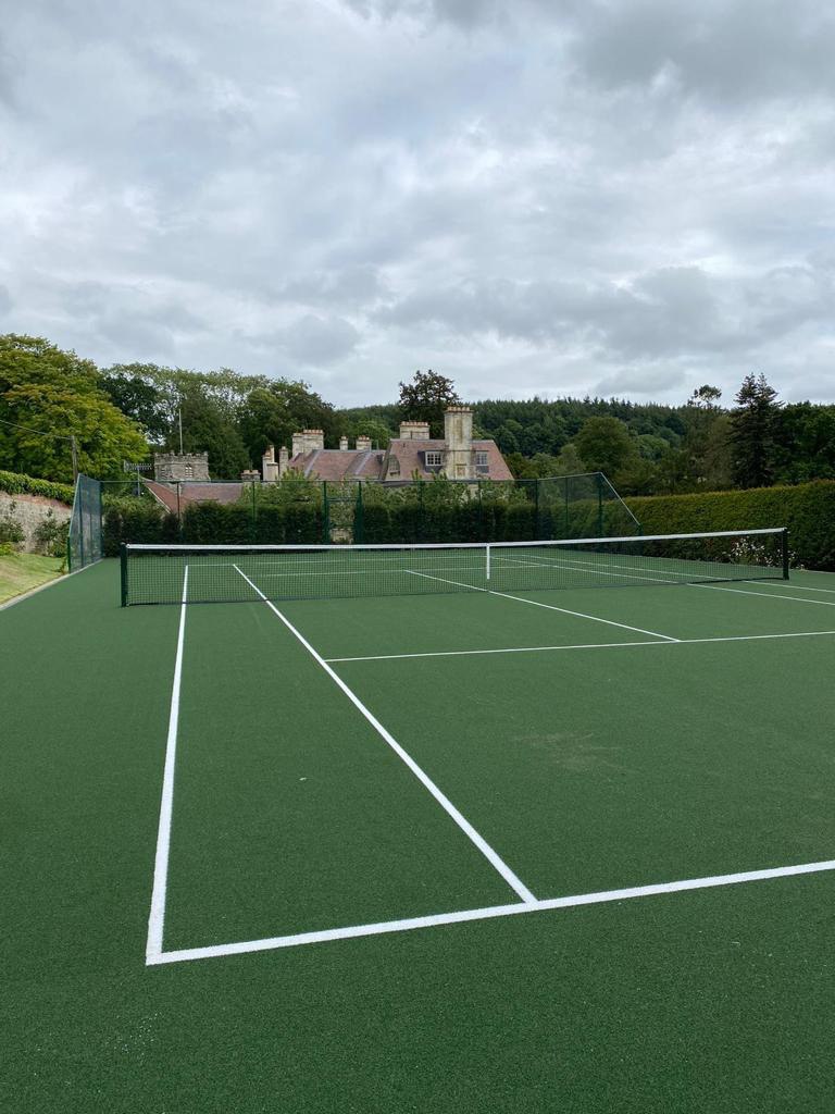 This is a photo of a newly installed tennis court, the court has a drop fence on both sides, one side is enclosed by a hedge. In the distance there is a very large county house and trees.