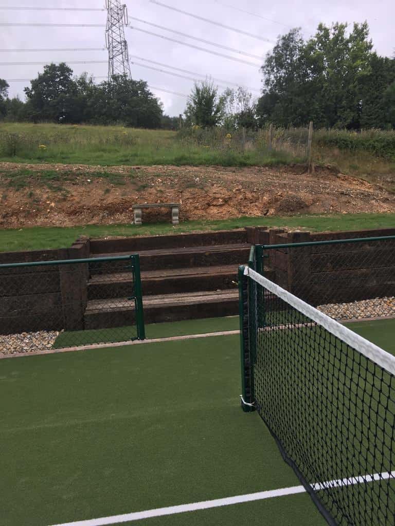 This is a photo of a newly installed tennis court, with a new green metal fence, and a gate by the service line. Behind the gate there are steps made out of wooden sleepers.