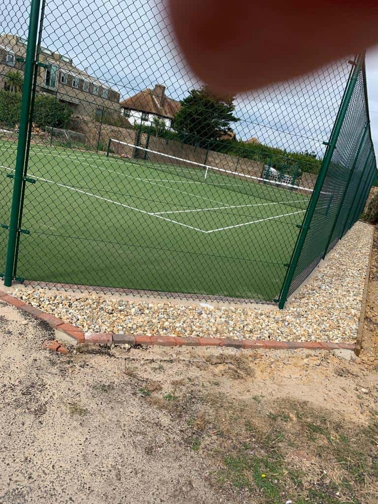 This is a photo of a new tennis court with buildings in the background. Around the edge, landscaping is required but the court has been finished.