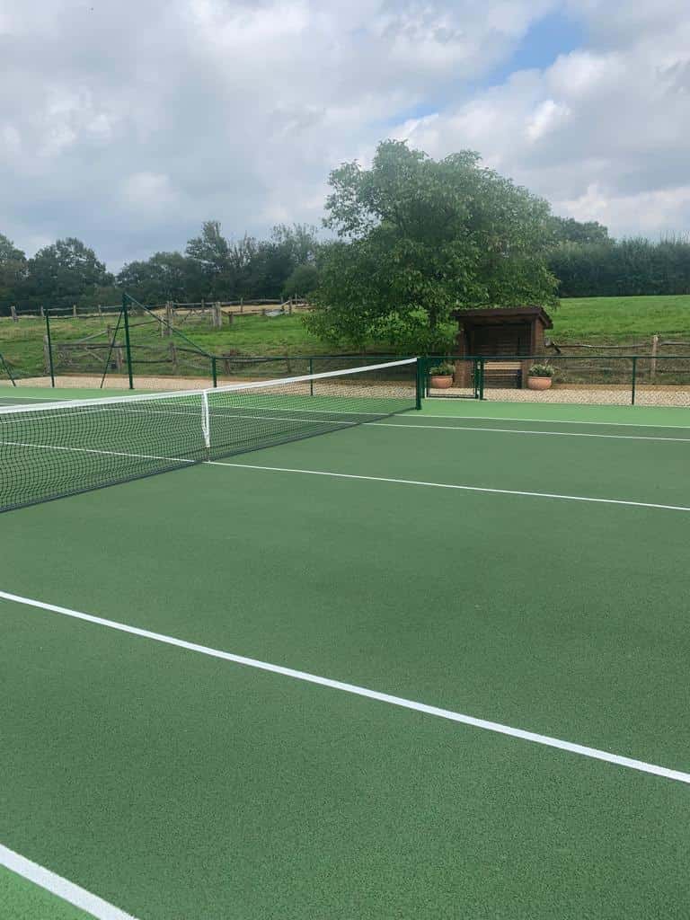 This is a photo of a newly constructed tennis court, which has been taken whilst standing on the court looking out to the side of it, where there is a seating area and a tree. The court has been freshly landscaped as well.