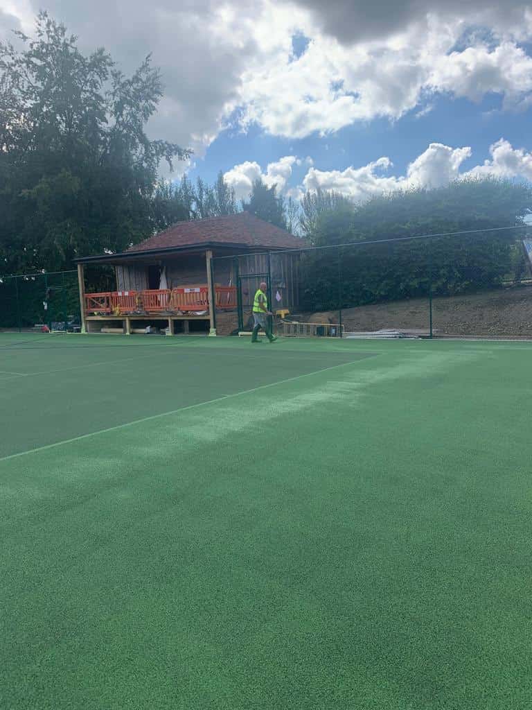 This ais a photo of a tennis court in the process of being resurfaced. There is also a viewing area located to the side of the court, and an operative working on the court