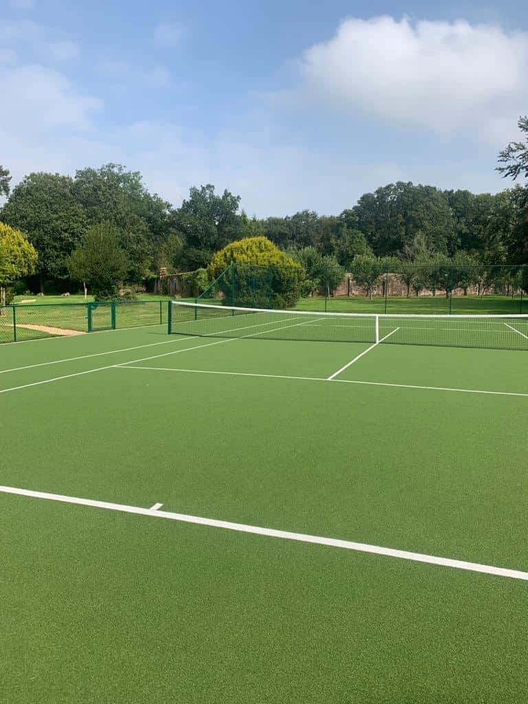 This is a photo of a newly installed tennis court taken from behind the serving line showing the full court, including the lines, fence and net. In the background there are trees and a brick wall.