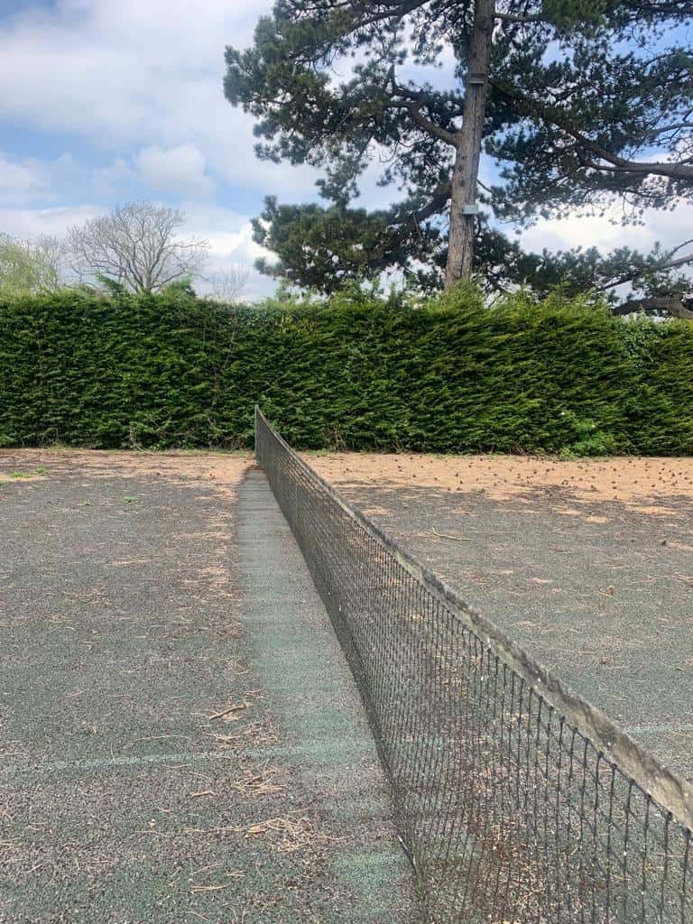 This is a photo of a derelict tennis court that needs resurfacing. The court is in very poor condition, and the net on the service line is very old and in poor condition. The tennis court requires full refurbishment.