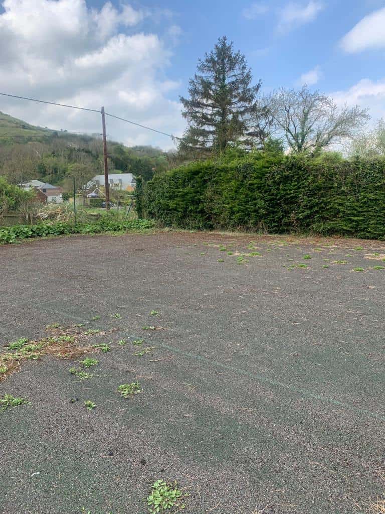 This is a photo of a derelict tennis court that needs resurfacing. The court is in very poor condition, there are weeds growing through the tennis court surface.