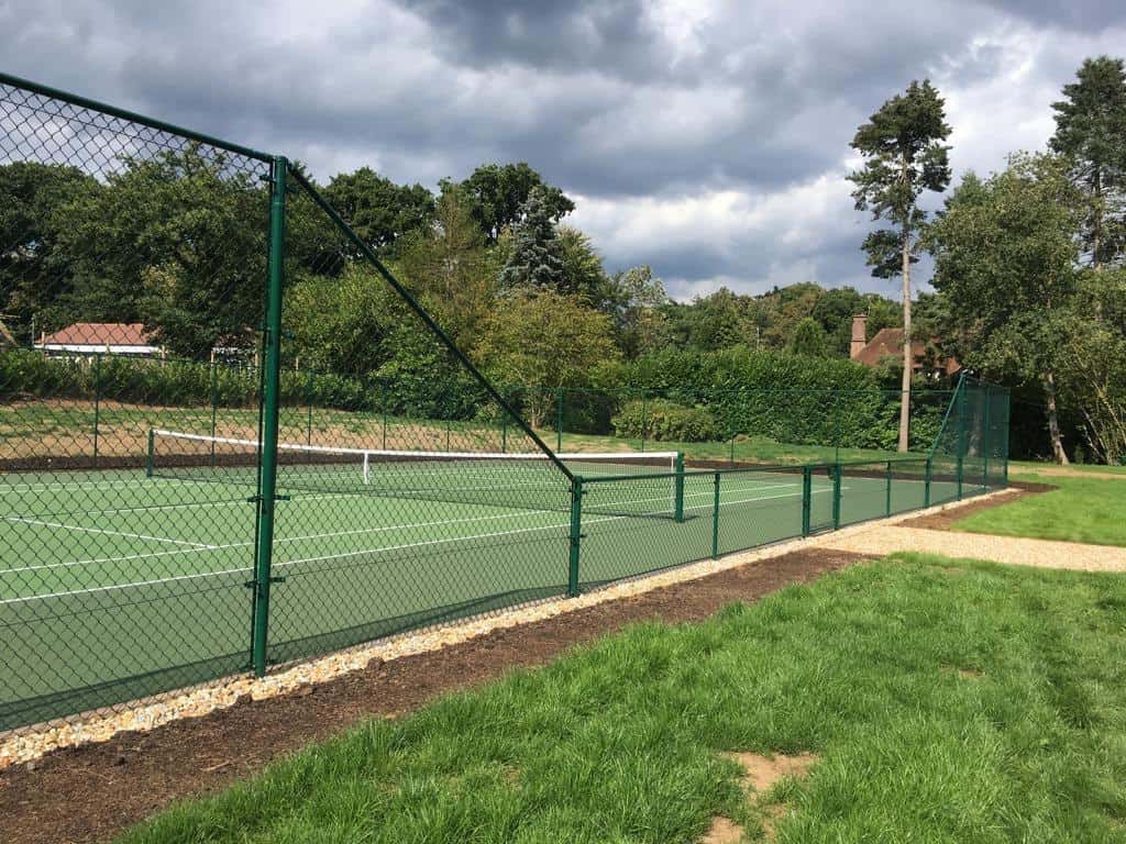 This is a photo of a new tennis court which is photo'd with a green fence surrounding it and a net in the middle. There is also a shingle path leading to the entrance of the court.