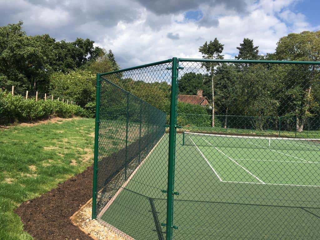 This is a photo of a newly installed tennis court the photo has been taken through the fencing and in the background there is a large country house.