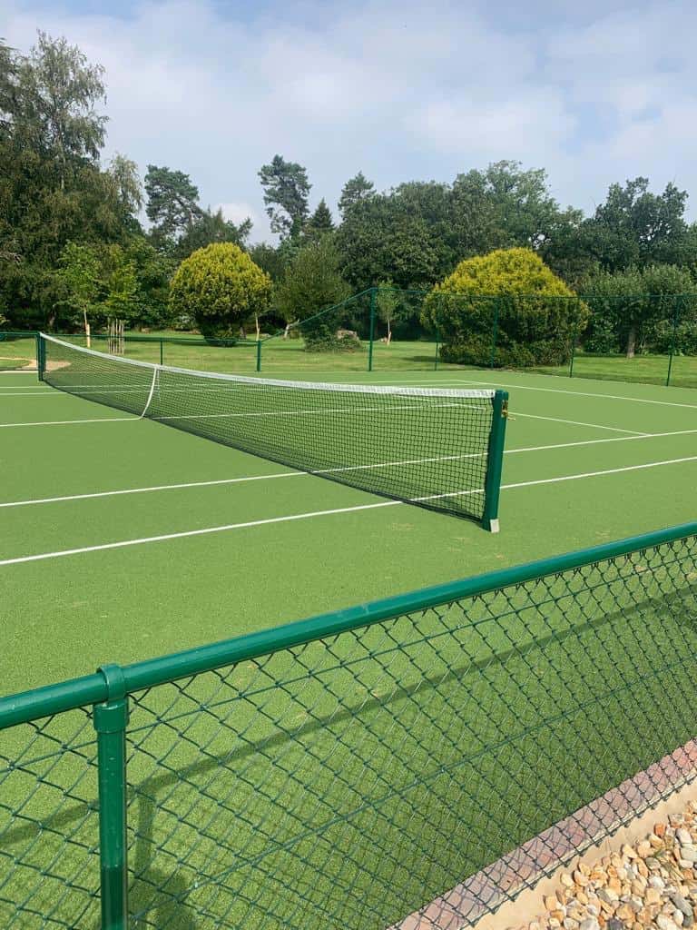 This is a photo of a newly installed tennis court taken from behind the serving line showing the full court, including the lines, fence and net. In the background there are trees and a brick wall.