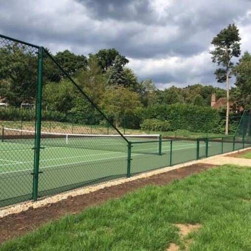 This is a photo of a new tennis court which is photo'd with a green fence surrounding it and a net in the middle. There is also a shingle path leading to the entrance of the court.